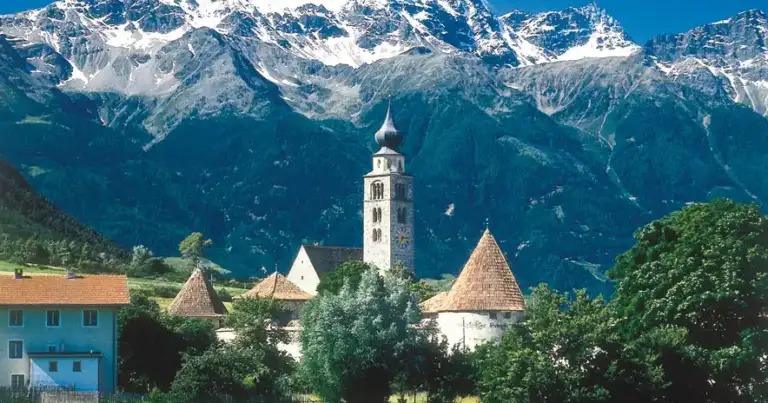 Pueblo alpino con iglesia blanca y torres contra montañas nevadas