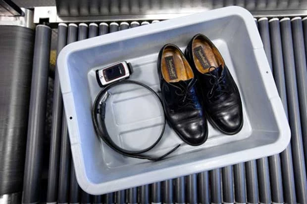 Bandeja gris con zapatos negros, un cinturón y un teléfono sobre la cinta de un control de seguridad
