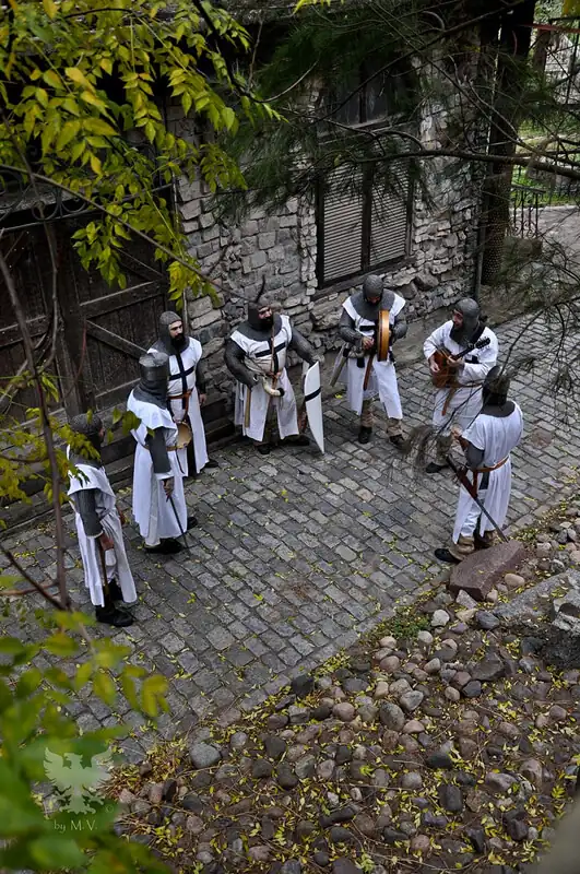 Personas en ropa medieval blanca caminando en plaza de piedra entre árboles