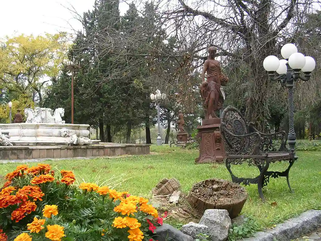 Parque con estatua de bronce, flores naranjas y farola entre árboles