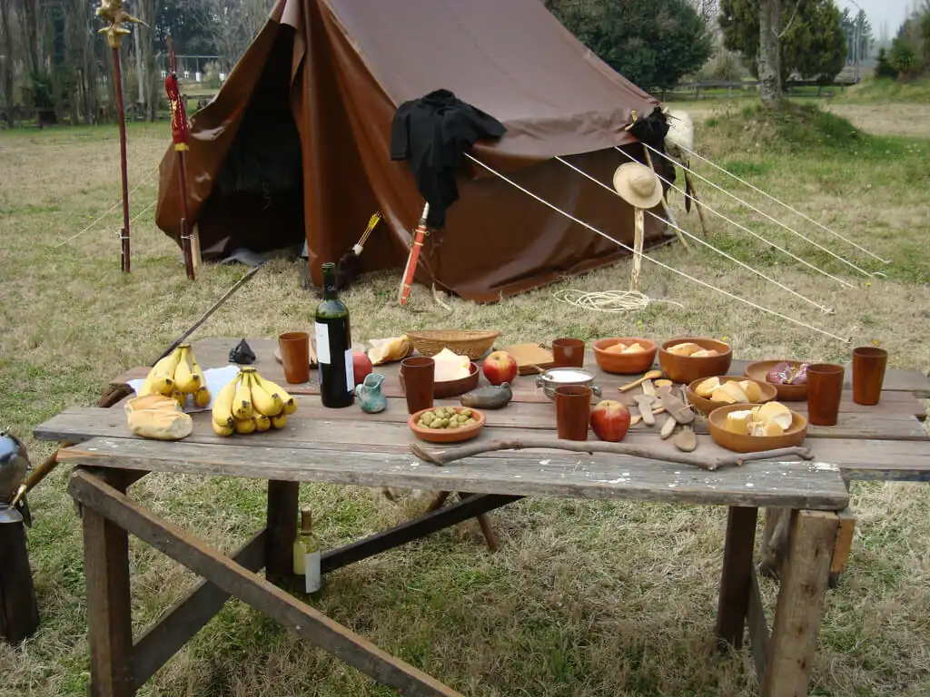 Mesa de madera con frutas, pan, jarras y cuencos de comida junto a una tienda de campaña marrón