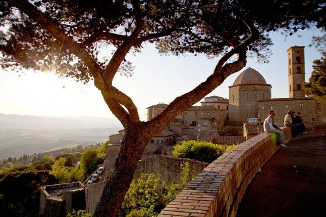 Mirador de piedra bajo un árbol con vistas a un pueblo de iglesia con cúpula y campanario, y al valle