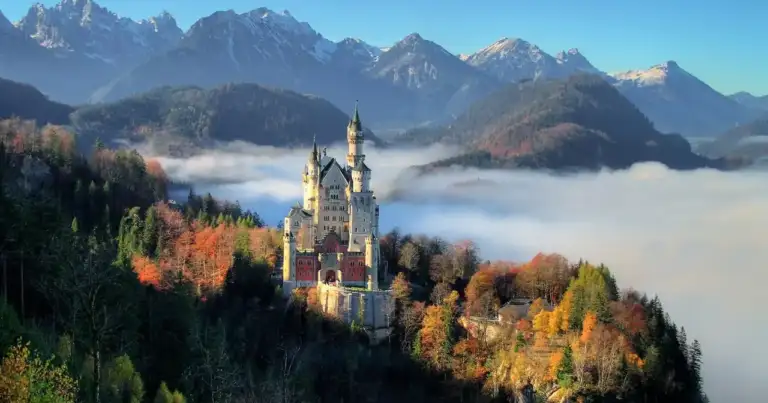 Castillo blanco rodeado de bosque otoñal y montañas nevadas