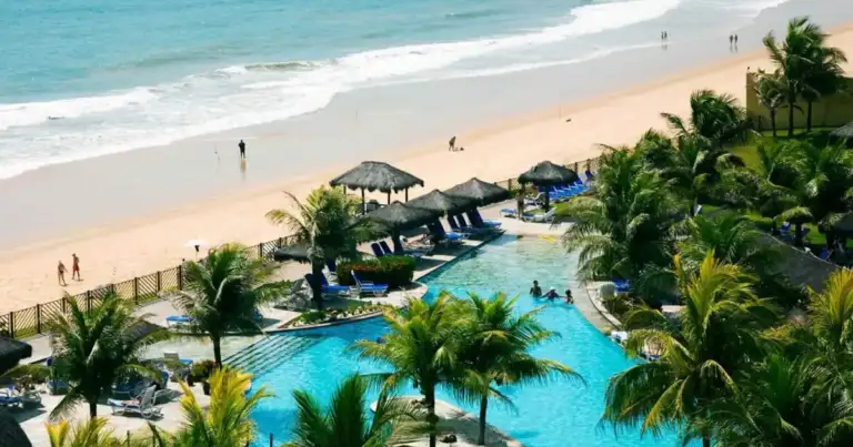 Piscina del resort frente a la playa en Brasil, rodeada de palmeras y sombrillas de paja.