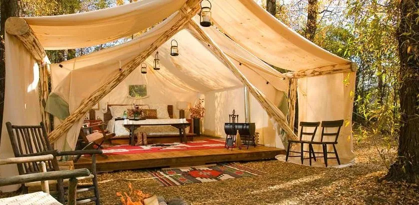 Carpa glamping de tela beige con cama interior, sillas y linternas entre árboles