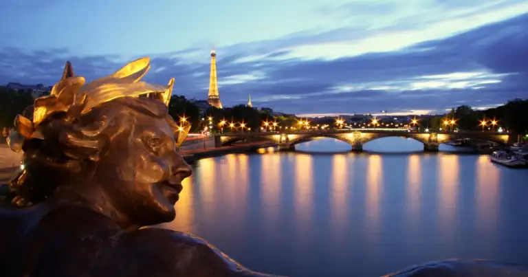 Estatua dorada iluminada junto a río al atardecer con puentes de ciudad reflejados