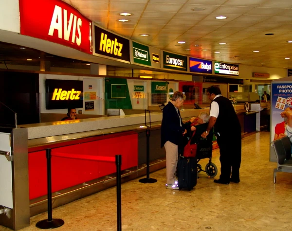 Mostrador de alquiler de autos en un aeropuerto, con logos de compañías como Avis, Hertz, Europcar y Budget. Dos personas en el mostrador reciben asistencia.
