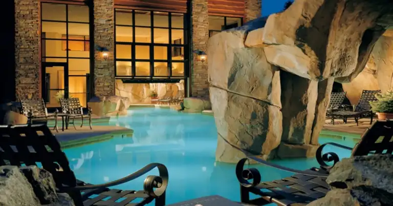 Piscina iluminada de noche rodeada de rocas y estructura de madera al fondo