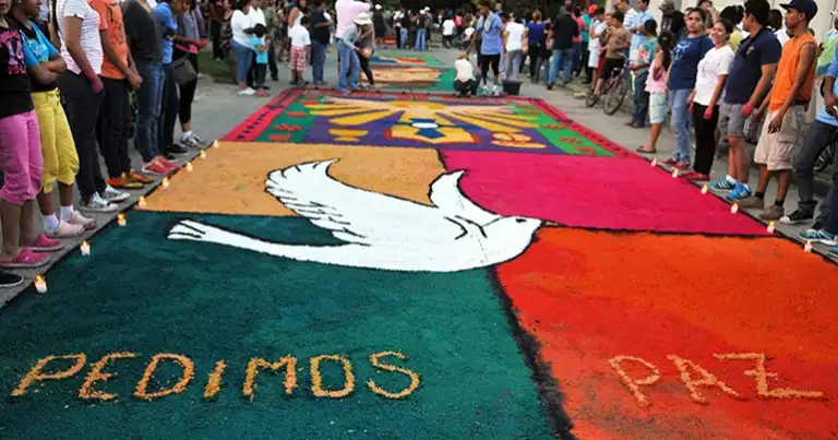Alfombra de aserrín multicolor en calle con imagen de paloma blanca y texto