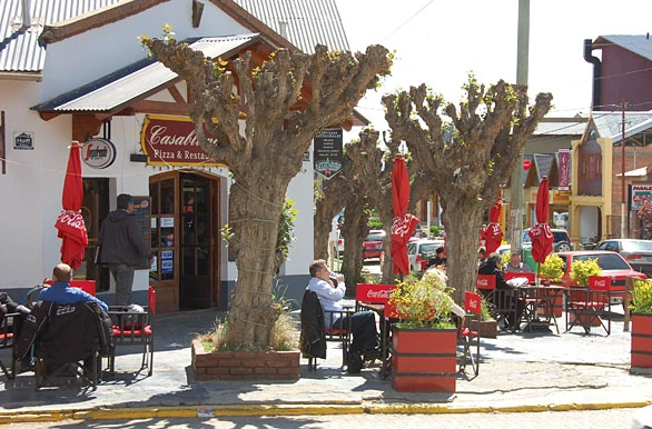 Terrazas con mesas y sombrillas rojas bajo árboles podados, frente a un local con el cartel Casablanca Pizza & Resto