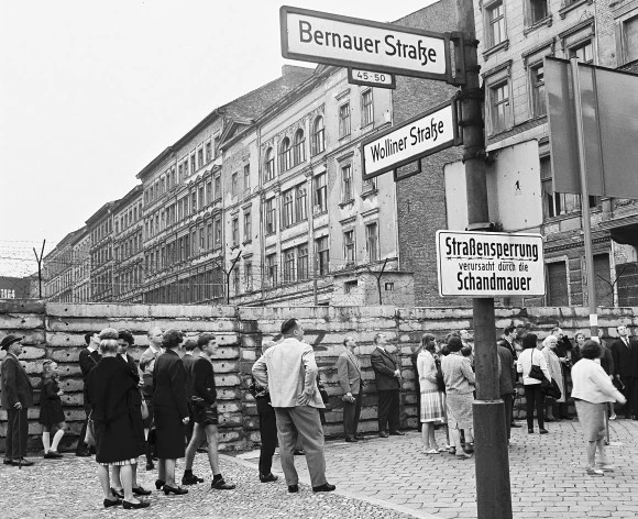 Fotografía histórica en B/N de calle con muro de ladrillos y carteles alemanes