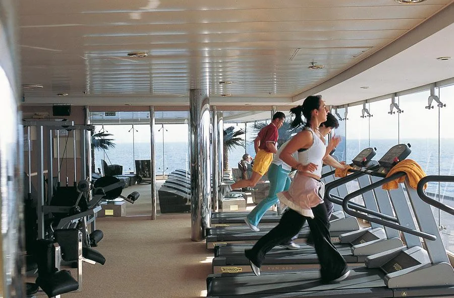 Personas haciendo ejercicio en gimnasio de crucero con máquinas y vista al océano