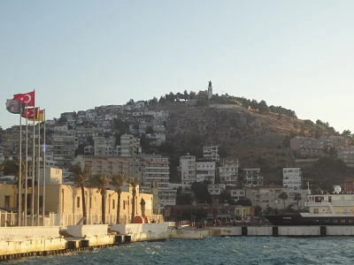 Ciudad costera de casas blancas sobre una colina, vista desde el mar, con banderas turcas junto al muelle