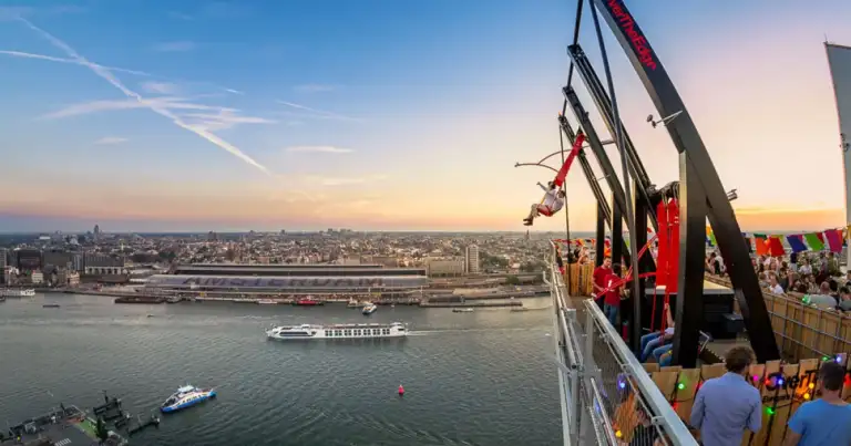 Atardecer sobre un río y la ciudad, con una atracción de plataforma elevada y gente en una terraza