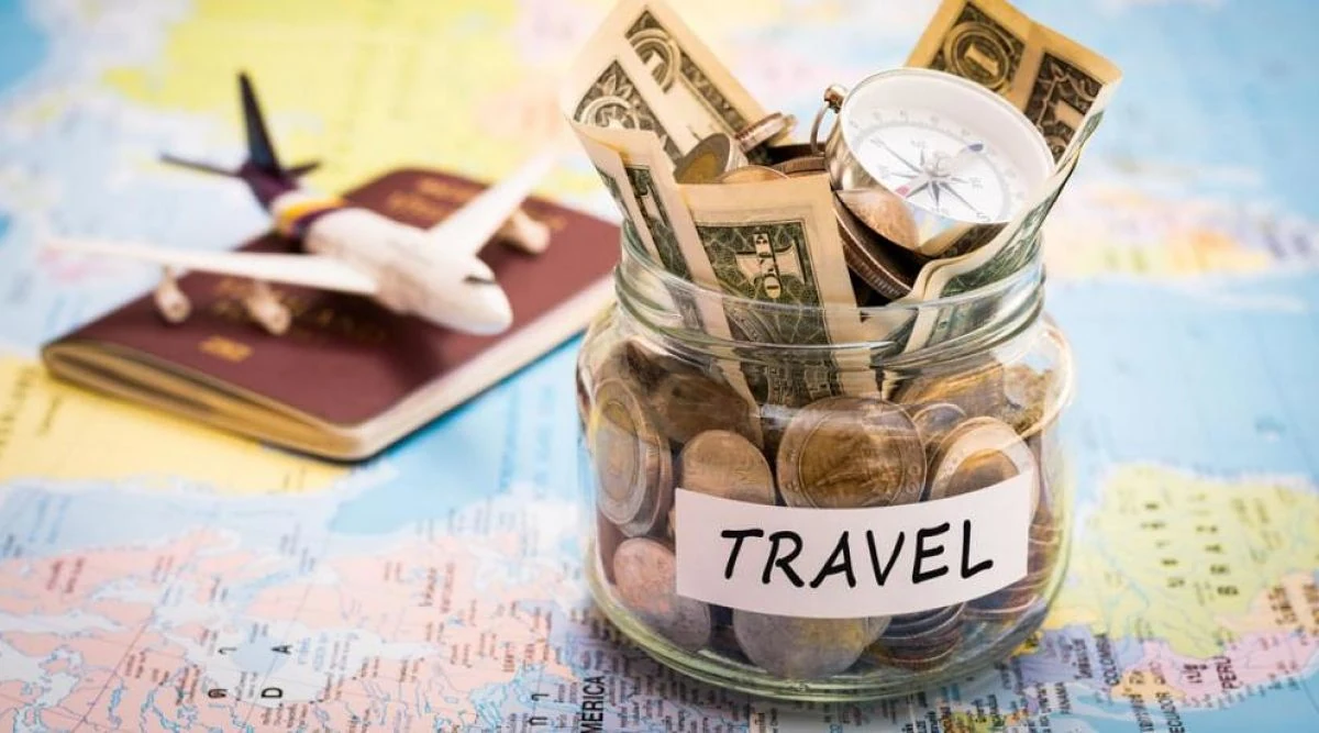 Frasco de vidrio con monedas, billetes y una brújula, rotulado «Travel», junto a un pasaporte y un avión de juguete