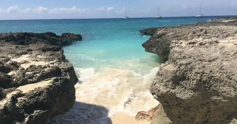 Playa de arena blanca con agua turquesa y rocas, barcos vela al fondo