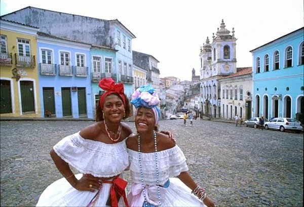 Dos mujeres con vestimenta tradicional brasileña en plaza colonial con edificios coloridos