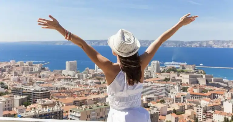 Mujer con brazos levantados y sombrero blanco viendo ciudad costera desde altura