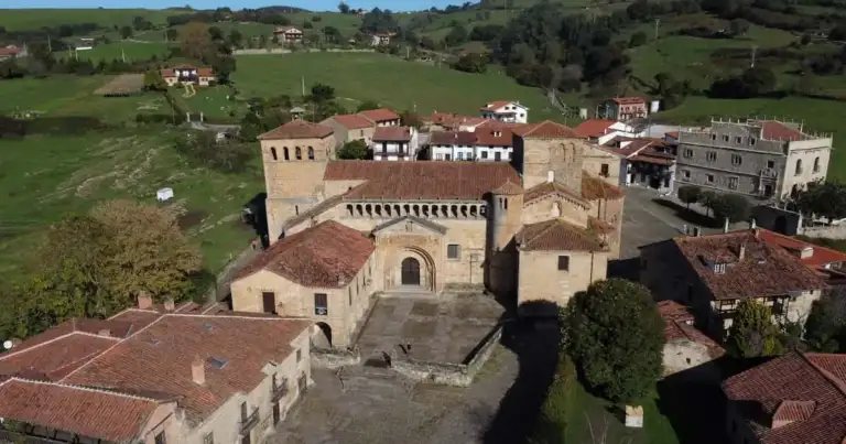 Vista aérea de la Colegiata de Santa Juliana en Santillana del Mar, rodeada de casas con techos rojos y prados verdes.