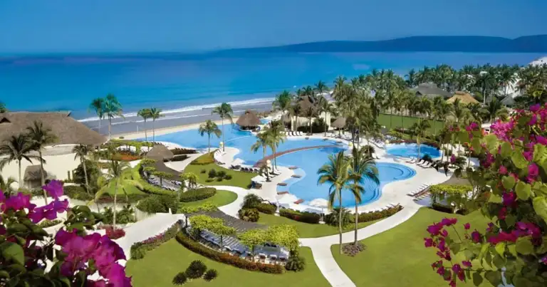 Complejo hotelero con piscina turquesa rodeado de flores magenta y playa