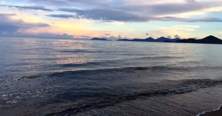 Playa de arena oscura al atardecer, con cerros lejanos y cielo nublado