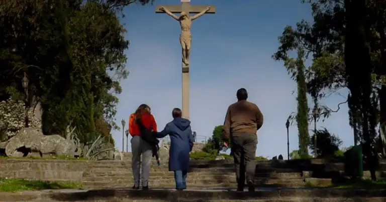 Tres personas contemplando cruz de madera en plataforma de piedra rodeada de vegetación