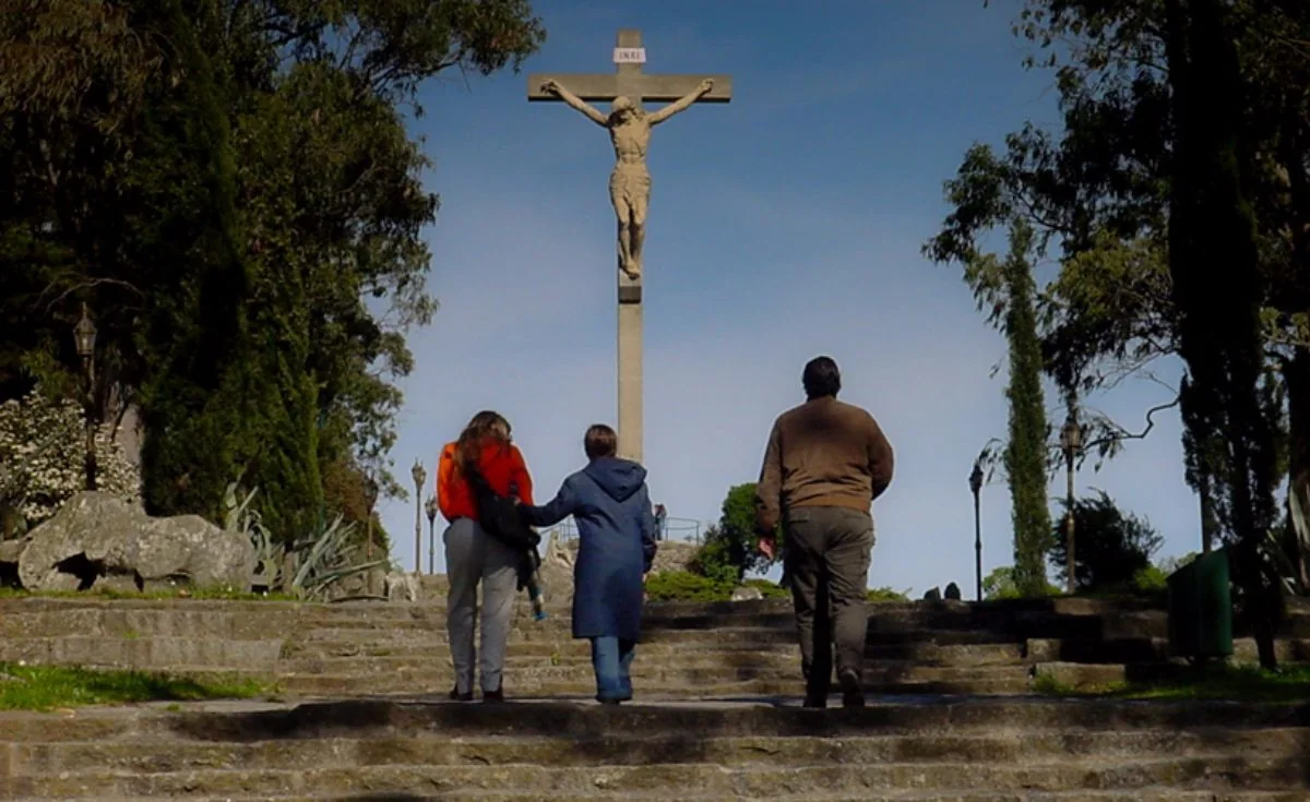 Tres personas contemplando cruz de madera en plataforma de piedra rodeada de vegetación