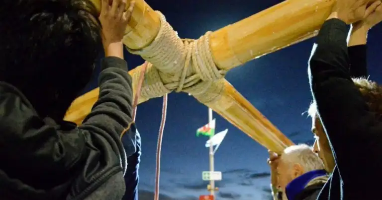 Personas sujetan una gran cruz de bambú atada con cuerdas, de noche