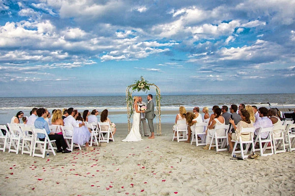 Ceremonia de boda en playa con novia, novio e invitados frente al océano bajo cielo azul