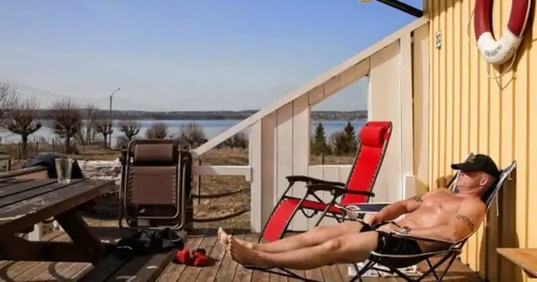 Hombre recostado en silla roja frente a lago bajo cielo azul
