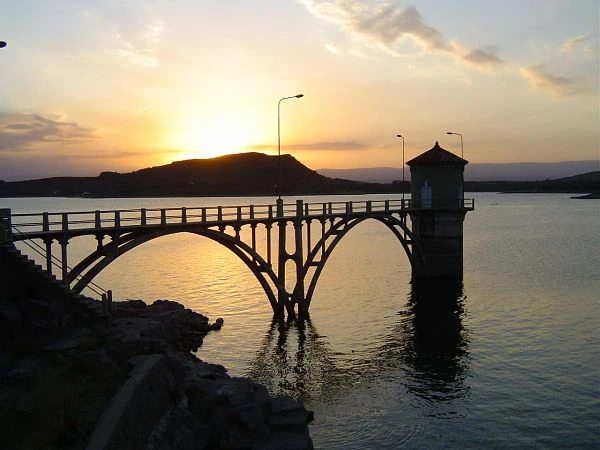 Puente de hierro sobre embalse al atardecer con montañas y agua reflejada