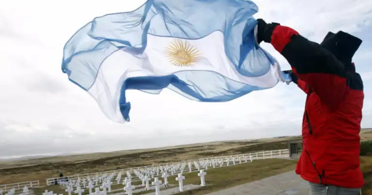 Persona con campera roja sostiene una bandera argentina al viento frente a un cementerio de cruces blancas