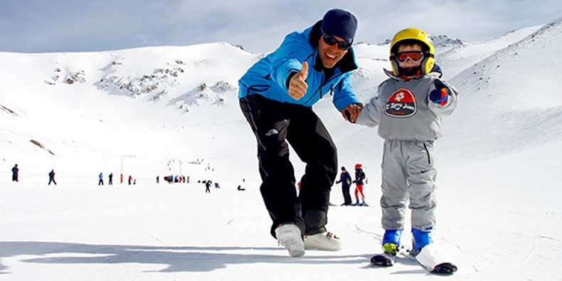 Adulto con chaqueta azul levanta el pulgar junto a un niño con casco amarillo que esquía en la nieve