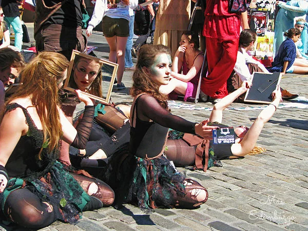 Artistas callejeras maquilladas y con trajes oscuros, sentadas en el empedrado, sostienen espejos y marcos
