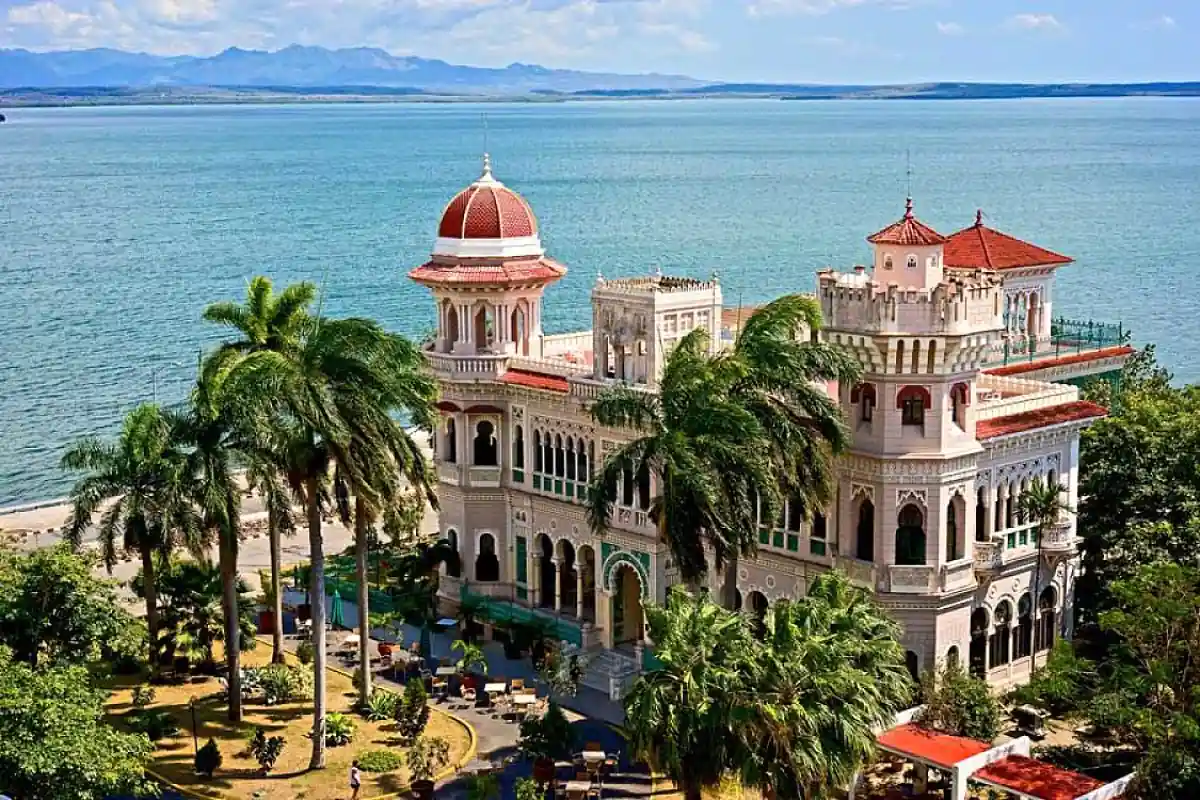 Palacio con cúpulas y torres rodeado de palmeras, junto a una bahía de aguas azules