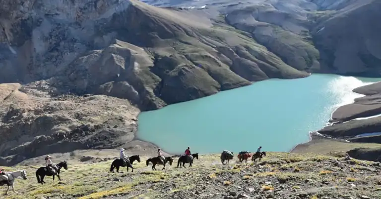Caballos y jinetes cruzando cordillera montañosa con lago turquesa