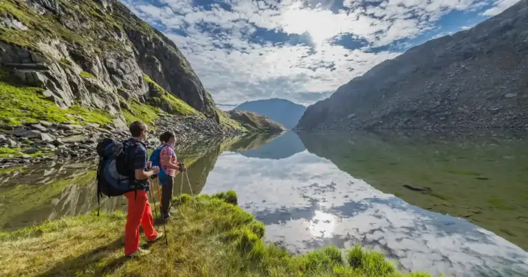 Dos senderistas en un acantilado verde mirando un lago alpino