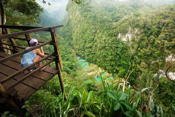 Viajero en plataforma de madera observando un profundo cañón con río turquesa y bosque tropical
