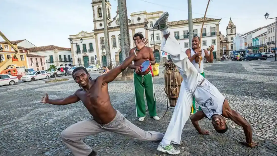 dos hombres de piel negra bailan en una plaza de bahía y una mujer con pantalón verde observa