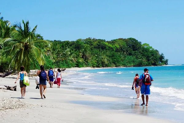 Turistas en playa de arena blanca con aguas turquesas, bosque tropical verde y olas suaves, Costa Rica