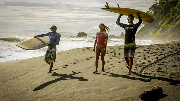 Tres surfistas en playa de arena con tablas de surf, olas al fondo y cielo nublado, Costa Rica