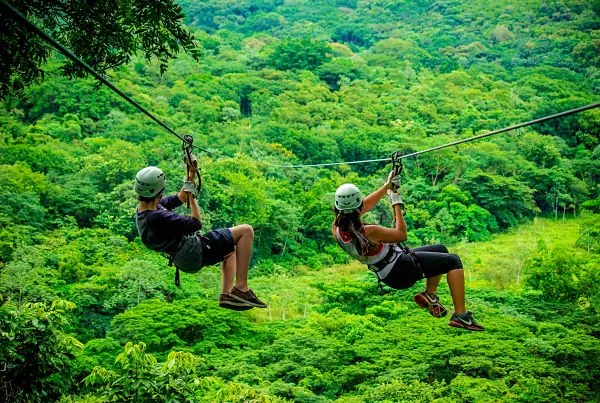 Dos personas con cascos y arneses volando en tirolesa sobre bosque tropical denso, Costa Rica