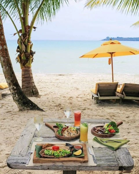 Mesas con ensaladas en playa tropical con sombrillas amarillas y palmera