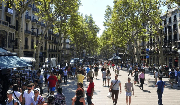 Paseo de Las Ramblas en Barcelona lleno de gente, con árboles frondosos, kioscos y edificios históricos a ambos lados, en un día soleado.
