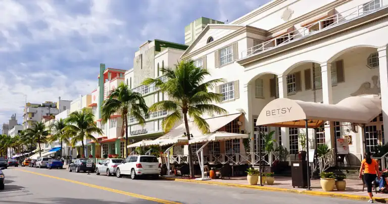 Calle Ocean Drive en Miami Beach con autos estacionados frente al hotel The Betsy, edificios art déco y palmeras bajo cielo parcialmente nublado.