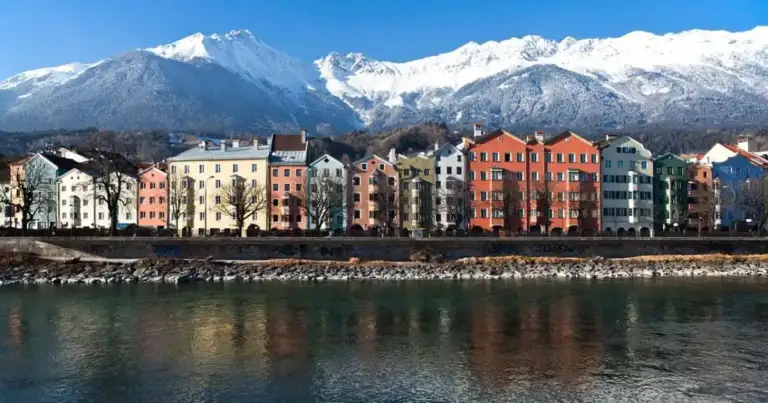 Edificios coloridos ribereños frente a un río con montañas nevadas al fondo