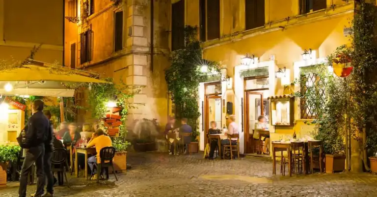 Plaza nocturna de un barrio europeo con edificios iluminados, terrazas y personas paseando