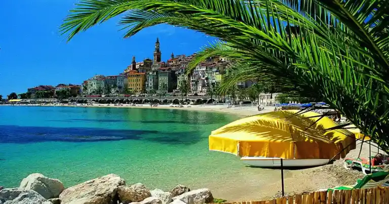 Playa mediterránea con sombrilla amarilla, rocas y pueblo costero al fondo