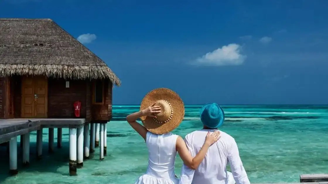 Pareja sentada en embarcadero tropical con bungalow sobre aguas turquesas