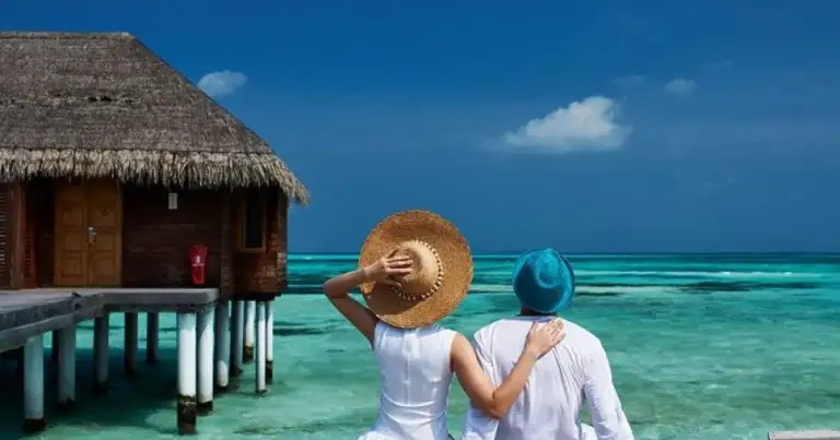 Pareja sentada en embarcadero tropical con bungalow sobre aguas turquesas